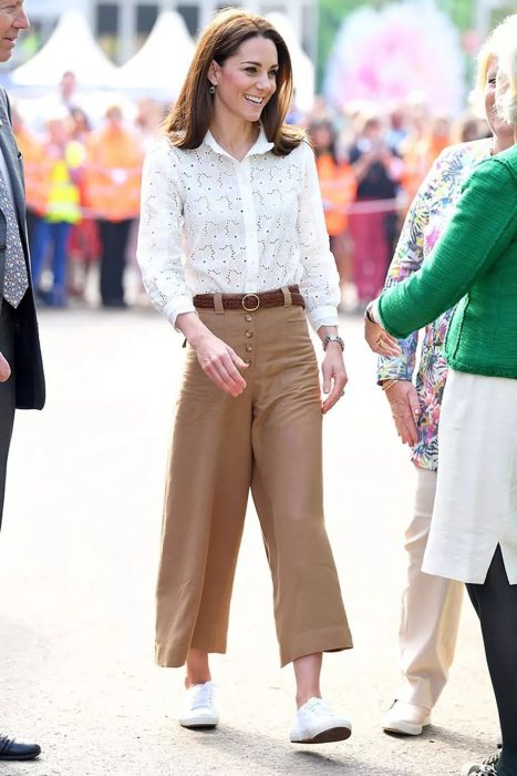 Kate Middleton vestindo culotes marrons, blusa branca e tênis branco 