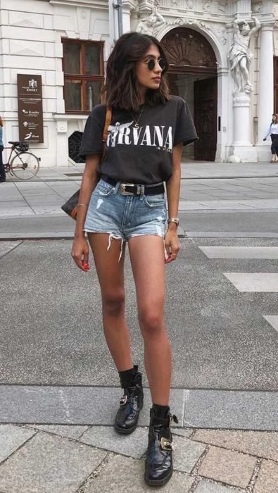 Menina com blusa preta nirvana e short jeans com cinto e botas engraxadas
