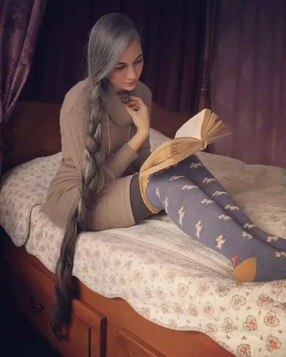 Mulher jovem de cabelos grisalhos, muito compridos e trançados para o lado, lendo um livro na cama em longas meias azuis com bonecos de cachorro