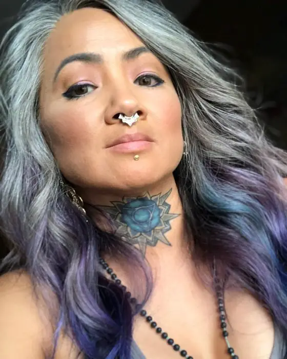Mulher com tatuagem de flor de lótus no pescoço, piercing no septo e cabelo cinza e roxo