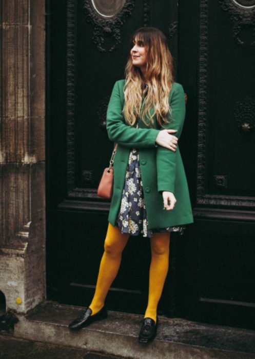 Trajes com meia-calça colorida;  garota posando em frente à porta, com meias amarelas, vestido florido e jaqueta verde