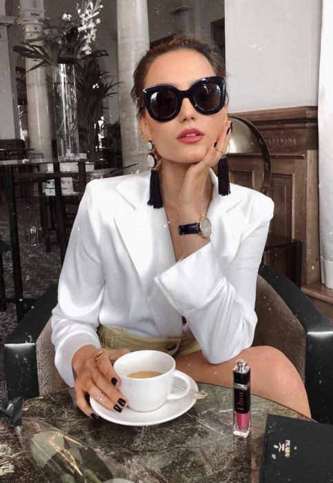 Conjunto com blusa branca;  mulher com óculos escuros, brincos de franja tomando um café