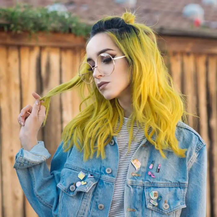 Menina com maquiagem profissional, óculos vintage redondos grandes, vestindo uma jaqueta jeans e alfinetes, tem cabelos amarelos com raízes pretas