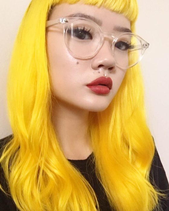 Menina asiática com lentes de aumento vintage grandes, piercing acima do lábio e cabelo longo e ondulado tingido de amarelo brilhante