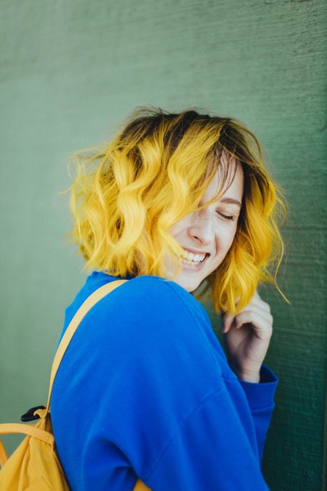 Menina sem maquiagem, sorridente, com suéter azul e cabelos ondulados e amarelo acima dos ombros