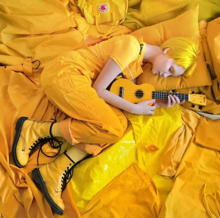 Menina com cabelo amarelo e corte pixie, com botas Dr. Martens e ukulele, rodeada de coisas amarelas