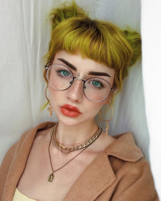Menina de olhos verdes, sardas, óculos com cabelos amarelos penteados com pãezinhos e piercing na ponte do nariz