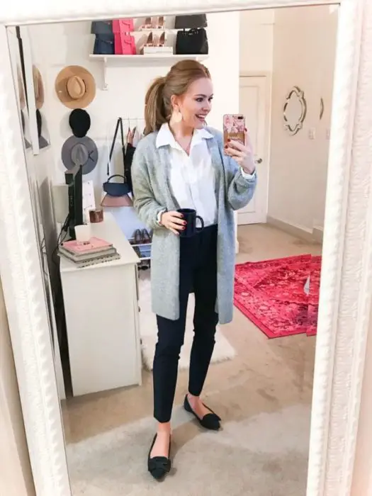Garota tira selfie em frente ao espelho com casaco de lã cinza, blusa branca e calça preta