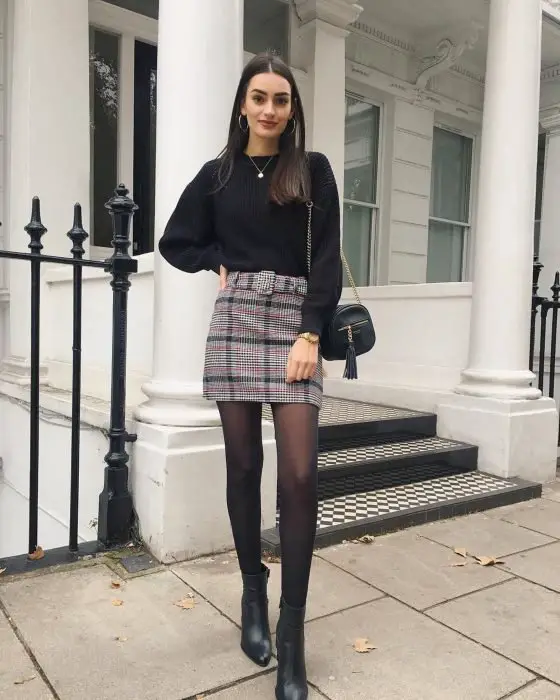 Garota fora de casa usando meia-calça preta, suéter preto e saia xadrez 
