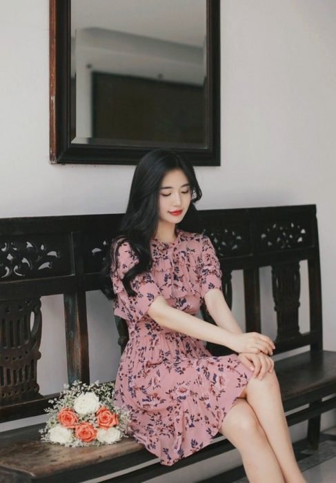 Menina coreana com cabelo preto ondulado, em um vestido florido na altura do joelho, sentada em uma banda e segurando um buquê de flores