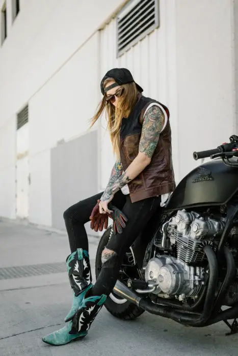 Parece moleca;  garota com roupa masculina com colete e botas de cowboy sentada em uma motocicleta estacionada