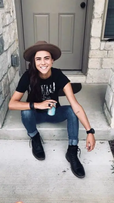 Parece moleca;  garota com roupa masculina, camiseta do Nirvana, jeans, botas pretas e chapéu, sorrindo sentada na varanda de uma casa