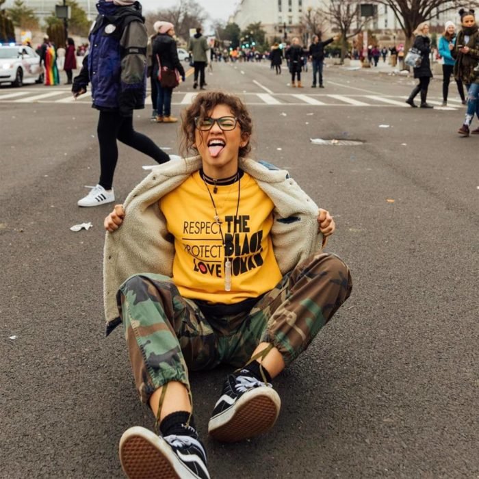Parece moleca;  menina com roupa masculina, Zendaya sentada na rua com calça militar, camisa amarela e jaqueta jeans com pele de carneiro, mostrando a língua