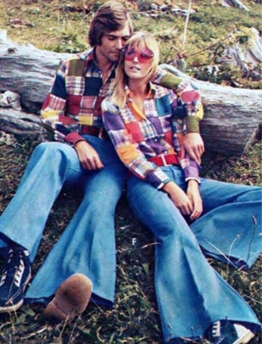 Moda feminina dos anos 70;  casal sentado em um tronco ao ar livre, camisa feita de retalhos de tecido, calça jeans larga e tênis preto e branco;  roupa retro