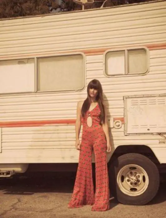 Moda feminina dos anos 70;  mulher na frente de um trailer com macacão vermelho com flores do decote até o umbigo, cabelo castanho longo e liso;  roupa retro