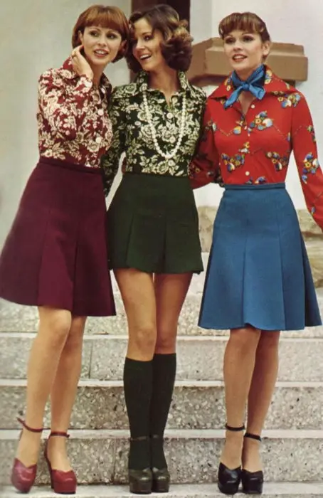Moda feminina dos anos 70;  três amigas se abraçando, em blusas florais cereja, verde e vermelha e saias cor de vinho e azuis até os joelhos, chinelos de salto alto e meias;  penteados e roupas retrô