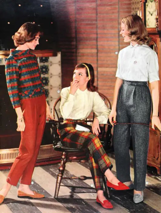 Moda feminina dos anos 70;  três amigos conversando e rindo;  calças cinza com quadrados vermelhos e amarelos, blusa branca e casaco estampado, sapatilhas laranja e cinza;  penteado retro e roupas