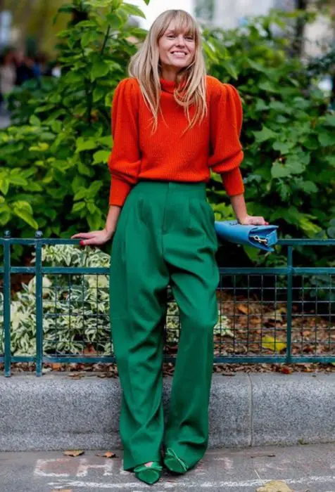 Looks com jeans largos;  mulher loira vestida com cores brilhantes e contrastantes, laranja e verde, com calças largas e suéter grande, bolsa azul