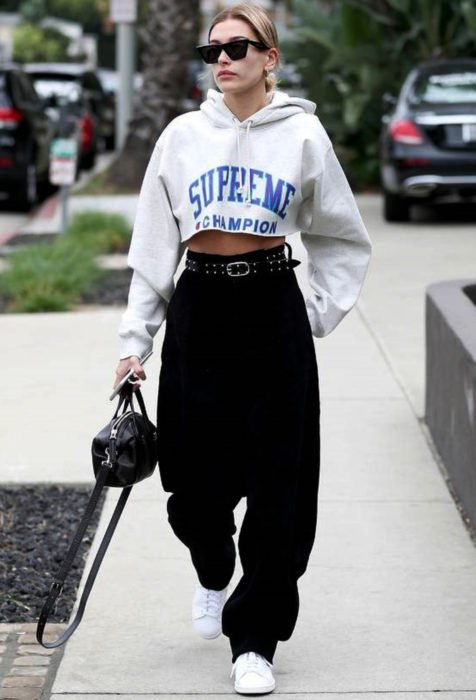 Looks com jeans largos;  Hailey Bieber andando na rua em um casaco com capuz crop grande, calças pretas largas com cinto de tachas e bolsa de mão