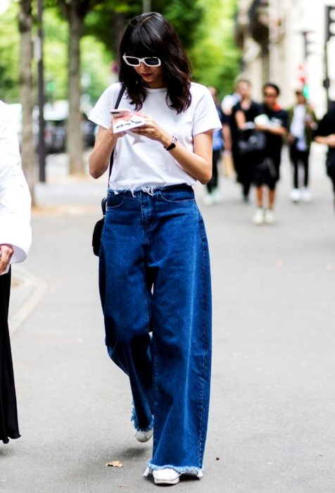 Looks com jeans largos;  mulher andando na rua olhando para o celular, vestindo uma blusa branca básica e calças largas