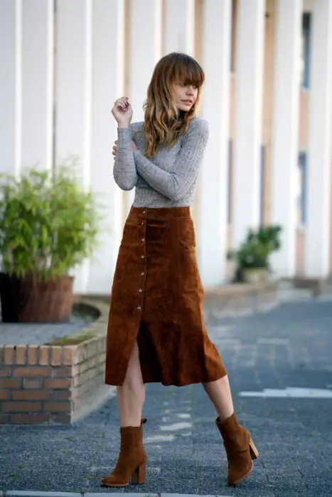 Menina vestindo uma saia marrom, botins e uma blusa cinza 