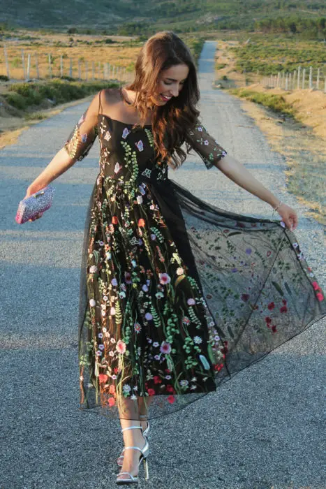 Idéias de vestidos para casamento em jardim ao ar livre;  garota andando na estrada feliz, em vestido de renda preta com flores e sandálias de salto fino, bolsa com glitter