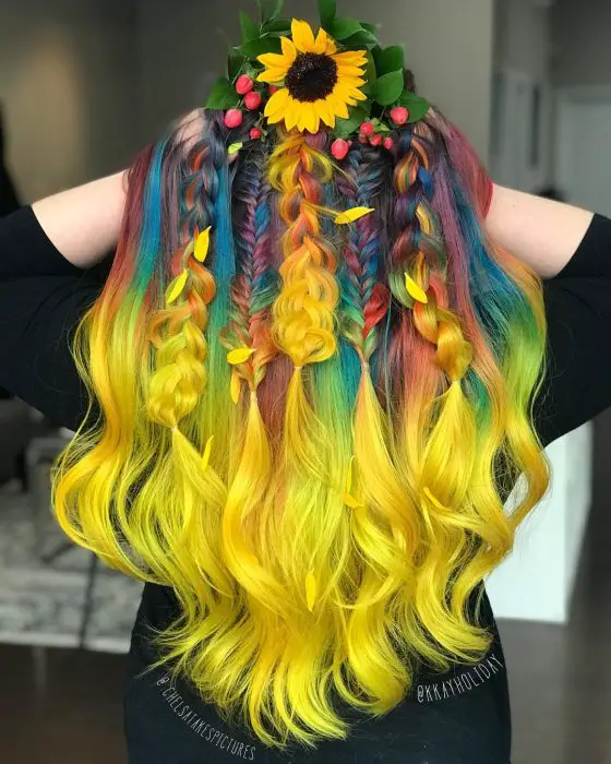 Menina com cabelo longo ondulado com tranças, cores do arco-íris, amarelo, verde, azul, laranja e roxo, usando uma coroa de flores de girassol