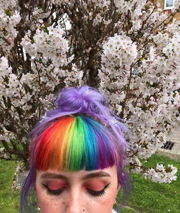 Menina com sardas e cabelo lilás com franjas vermelhas, laranja, amarelas, verdes, azuis e roxas em frente a uma árvore com flores brancas