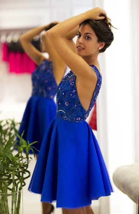 Menina de cabelos castanhos em frente a um espelho em um vestido de festa curto azul royal com enfeites e strass na parte superior e sem mangas