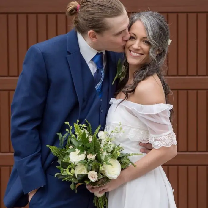 Casal de casais se abraçando, mulher com cabelos grisalhos, vestido de noiva branco e buquê de flores brancas, homem de terno azul com cabelo loiro, preso por um rabo de cavalo.