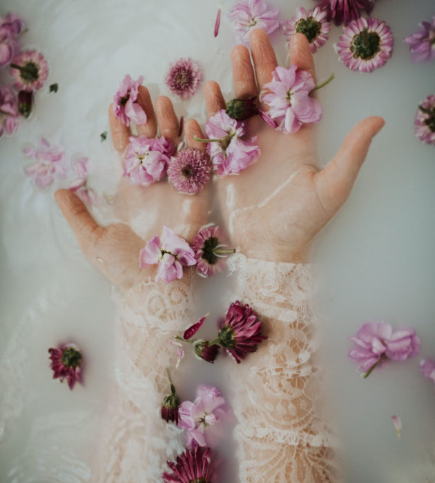 Banho de leite;  fotografia das mãos da mulher na água com leite e flores mroada