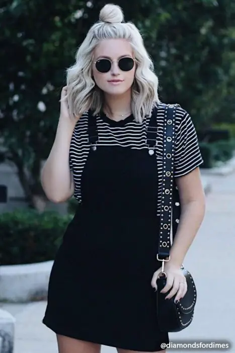 Garota em pé na rua usando um vestido avental preto e uma blusa preta com linhas brancas 