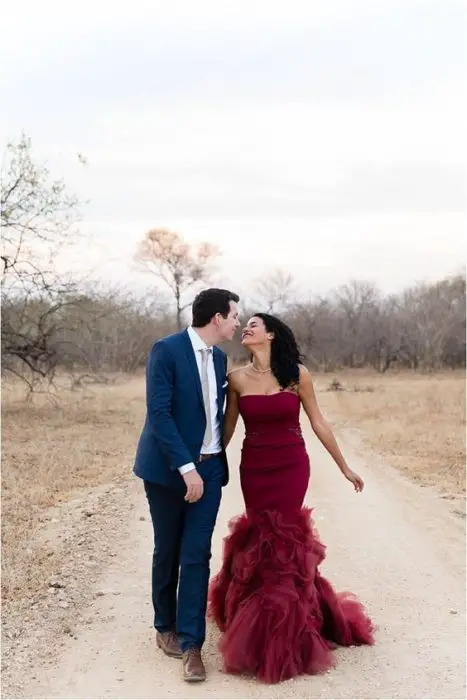 Um casal de noivos caminha por um campo amarelado, ela usa um vestido vermelho queimado