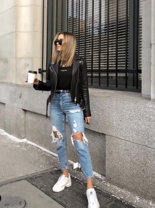 Menina parada no meio da rua segurando um café enquanto usava jeans rasgados, jaqueta preta e blusa e tênis de linha branca 