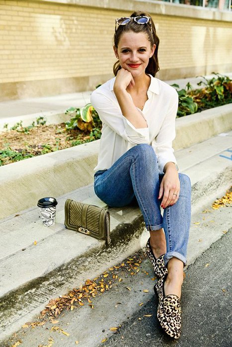 Mulher de blusa branca, jeans e sapatos com estampa animal sentada no banco com a bolsa de lado