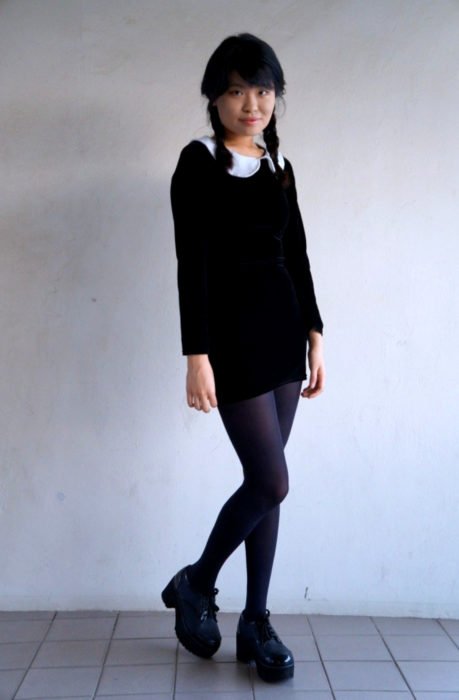 Menina asiática vestida de Merlina Adams;  vestido preto curto com gola branca, sapatos plataforma e penteado trançado