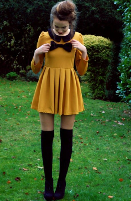 Menina vestida de Merlina Adams;  vestido amarelo mostarda com laço preto, meias, botinhas, penteado com coque alto