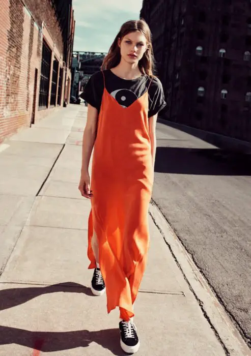 Looks com vestidos e blusas básicas;  mulher andando na rua com vestido maxi laranja, blusa com estampa yin e yang, tênis preto e cabelo comprido e desgrenhado