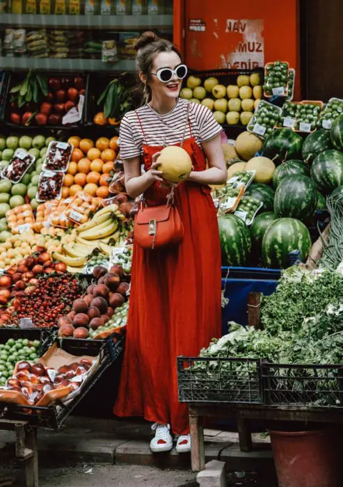 Looks com vestidos e blusas básicas;  mulher de verdureiro no meio de frutas, com coque alto, óculos de sol branco, maxi vestido vermelho, blusa listrada, tênis