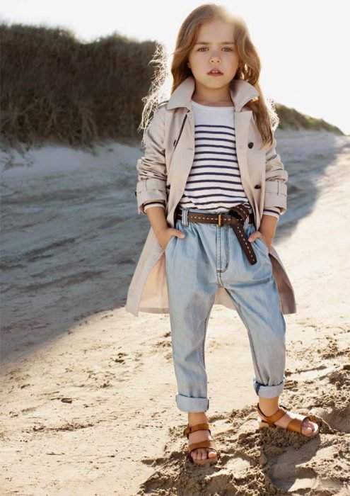 Menina fashionista vestida com jaqueta, calça jeans e sandálias em pé de frente para o mar 