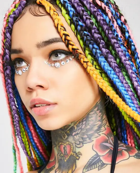 Looks com tranças africanas;  mulher de olhos cinzentos, com cabelo arco-íris, laranja, verde, azul, roxo e rosa, com tatuagem de coruja, rosa e coração no pescoço, com pedras nos olhos