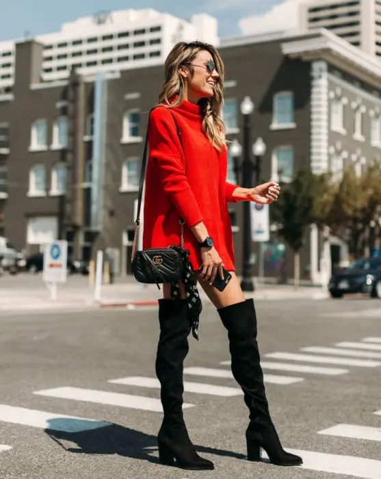 Mulher loira na rua, sorrindo e vestindo um suéter vermelho enorme como vestido de outono com botas pretas e longas de camurça