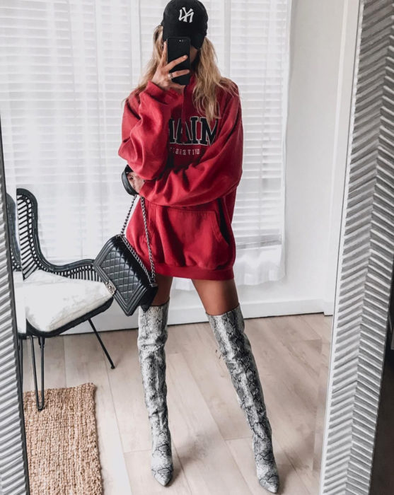 Mulher loira tirando uma selfie em frente ao espelho, vestindo um moletom vermelho enorme como um vestido de outono, com botas longas de cobra cinza com estampa animal