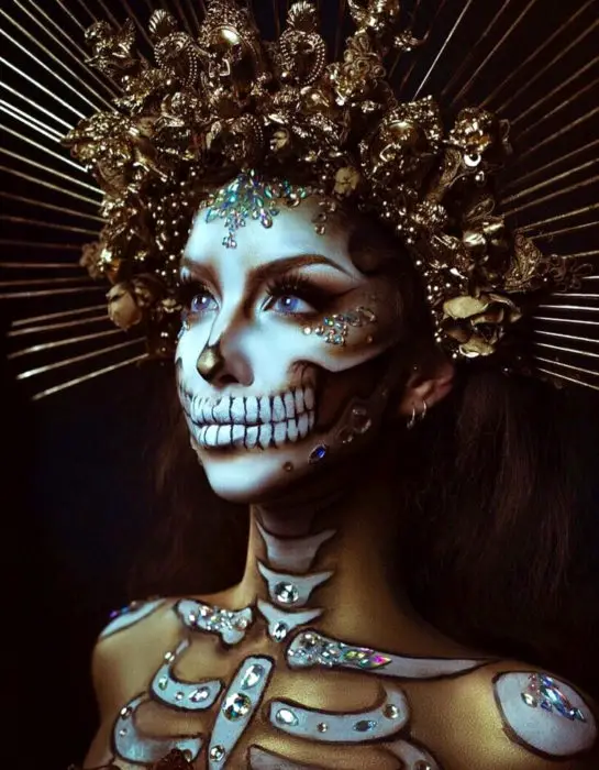 A maquiagem moderna de Catrina para o Dia dos Mortos;  mulher de olhos azuis com coroa dourada e pedras swarovski no rosto