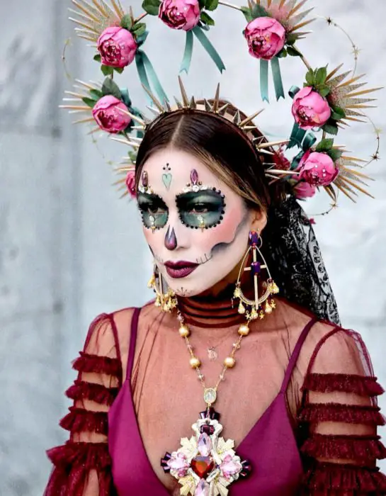 Maquiagem Modern Day of the Dead Catrina, com tiara dourada com pontas e coroa de estrelas e flores rosa, maxi colar e brincos em cruz
