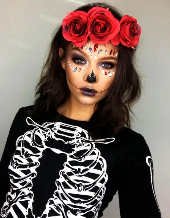 Maquiagem Modern Day of the Dead Catrina, sombra roxa e preta, com pedras swarovski no rosto, com blusa com estampa de caveira, coroa de flor vermelha