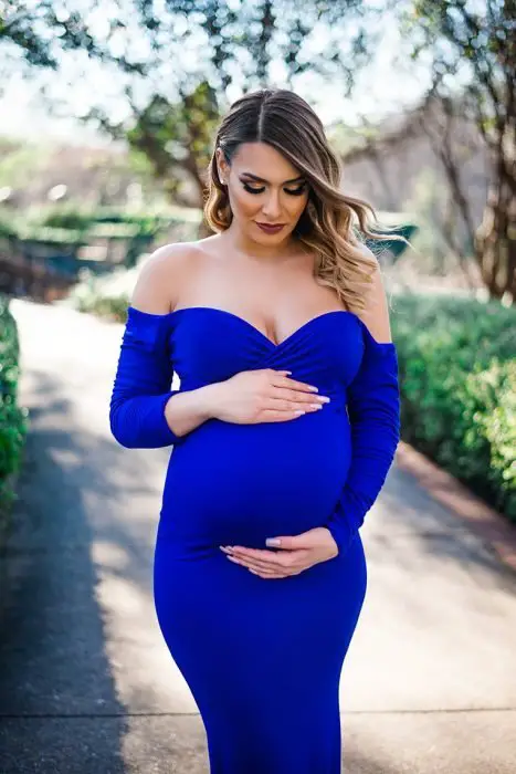 Vestido para chá de bebê, grávida, com cabelos castanhos com mechas californianas, usando vestido azul royal sem ombros, agarra a barriga