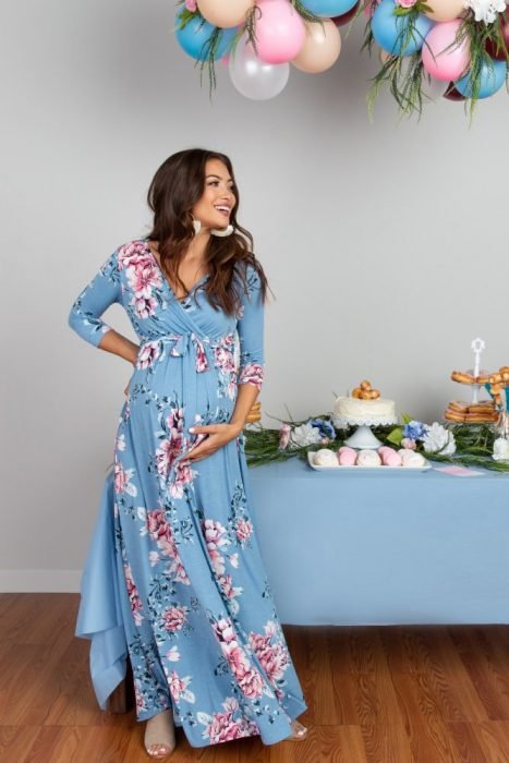 Mulher grávida de cabelos castanhos, usando um vestido azul, comprido e folgado, flor rosa, comemorando o chá de bebê
