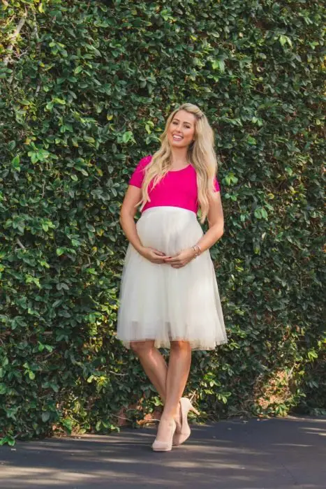 Vestidos de chá de bebê, menina loira com vestido rosa e branco grávida com tule, chinelos de salto baixo e posando em frente a uma parede com trepadeira