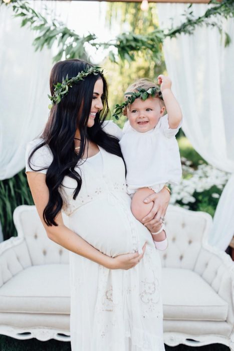 Vestidos para o chá de bebê, gestante carregando seu bebê com roupas combinando, mãe com vestido branco e coroa de flores e bebê com manto branco
